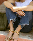 Person sitting on a ledge with a woven bag next to them, wearing gold sandals and a white top.