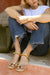 Person sitting on a ledge with a woven bag next to them, wearing gold sandals and a white top.
