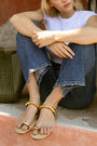 Person sitting on a ledge with a woven bag next to them, wearing gold sandals and a white top.