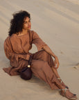 Woman in a brown dress sitting on sand with a textured background, wearing snake skin thong sandals